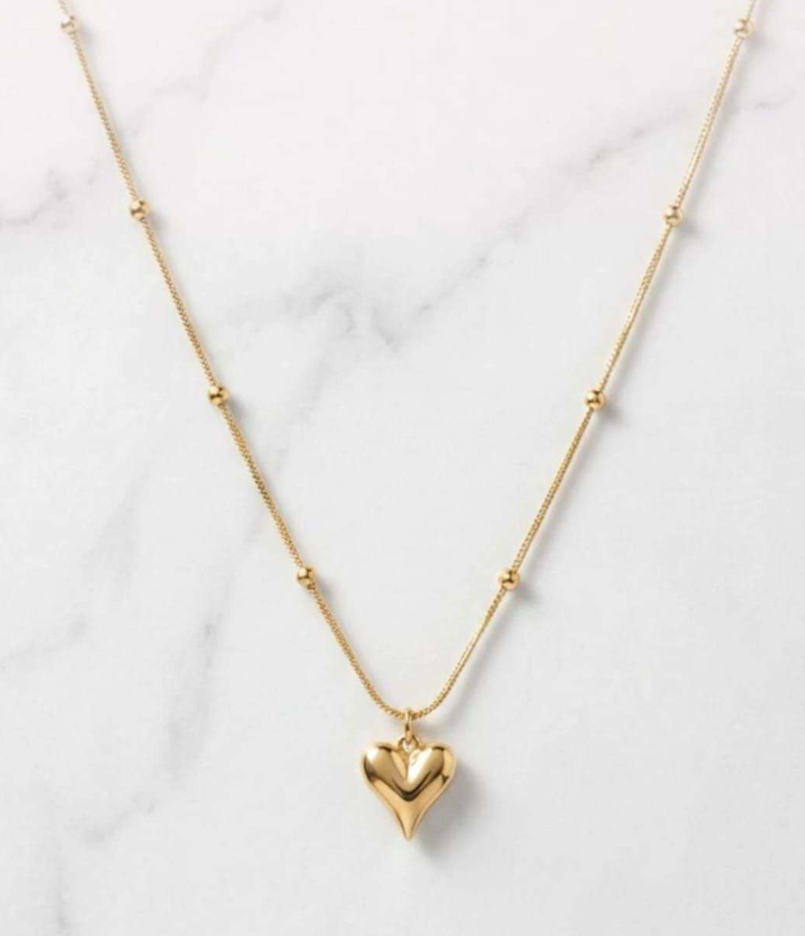 Gold-tone puffed heart pendant on a beaded satellite chain necklace laid on marble