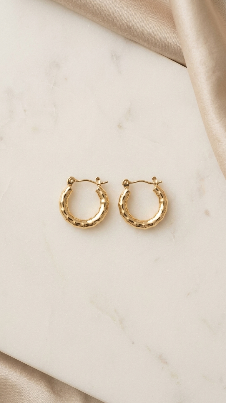 A pair of gold-tone hexagonal huggie hoop earrings with a segmented bamboo-style texture, resting on a shimmering ivory fabric background.