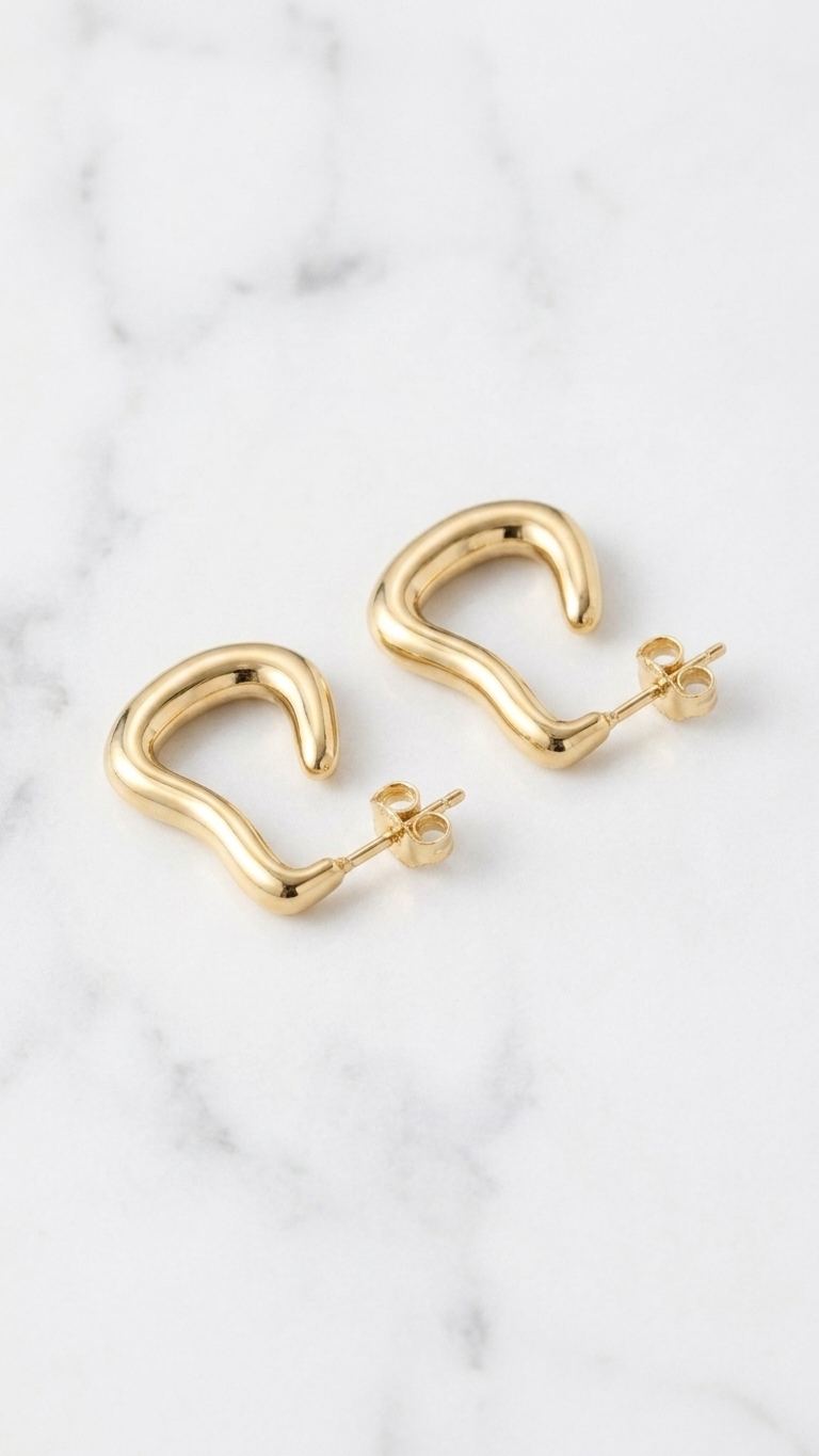 A pair of gold-toned J-hoop earrings with an irregular, wavy organic shape and a high-shine finish, shown on a white marble surface.