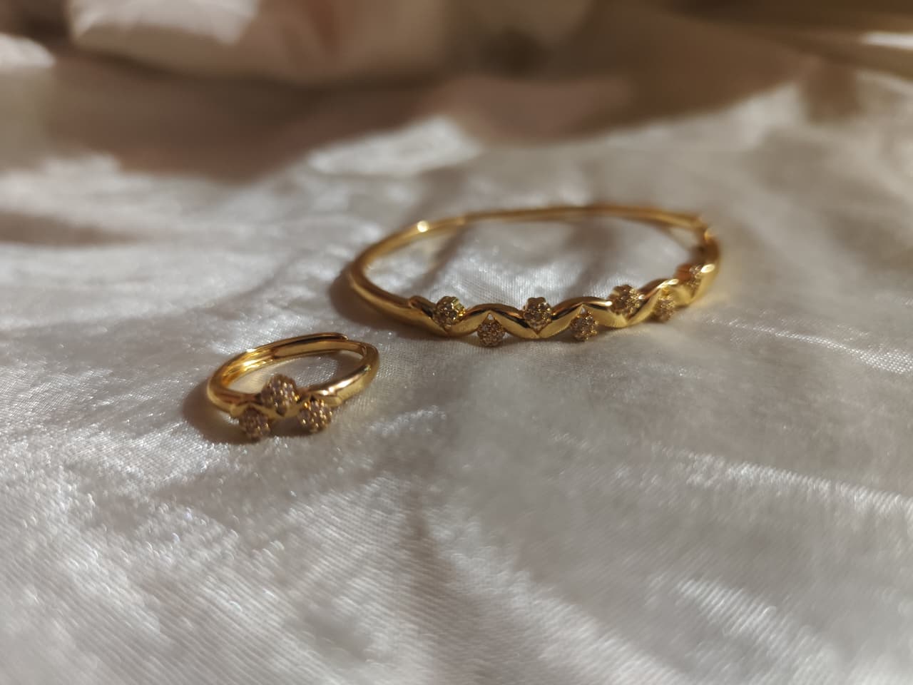 A matching gold-tone jewelry set consisting of a wavy bangle and a ring, both featuring small crystal clusters, displayed on a soft white velvet surface.