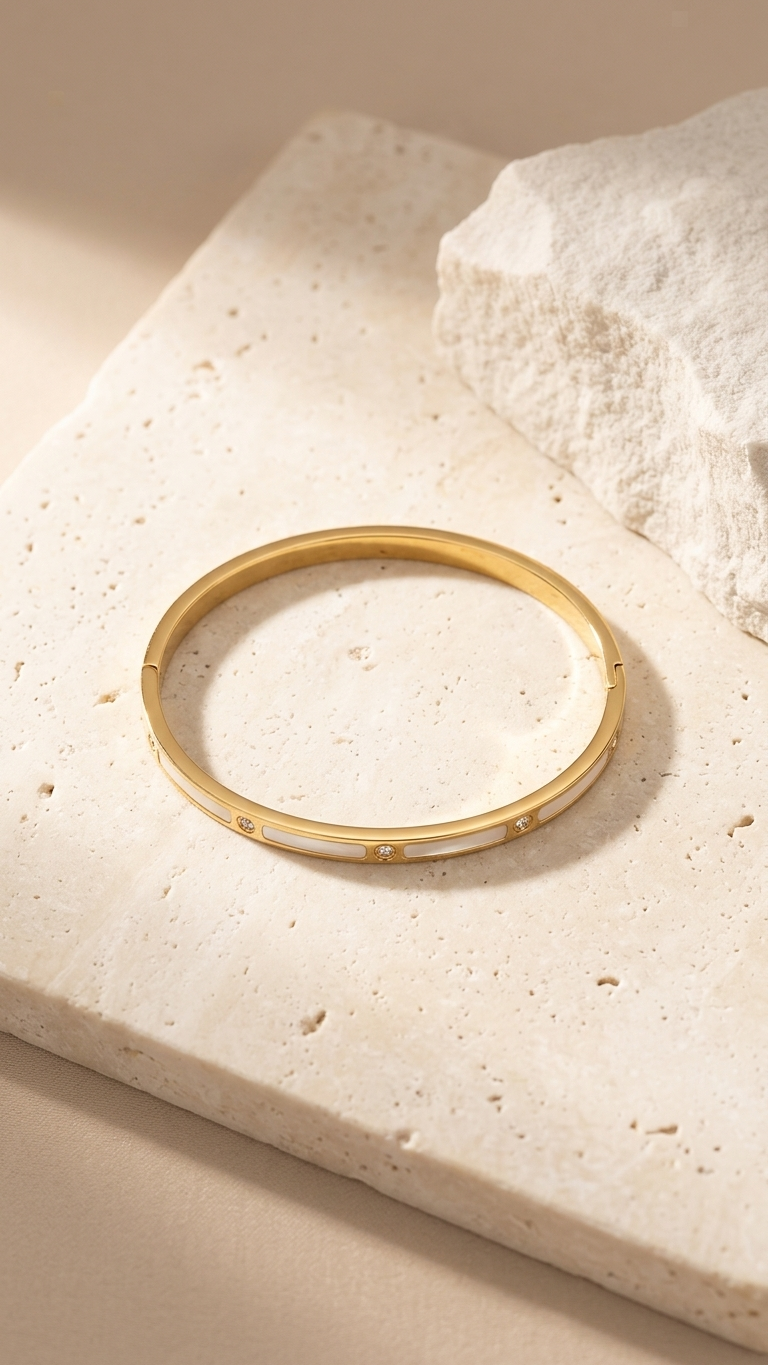 A gold-tone bangle with white rectangular inlays and small round crystals between segments, resting on a textured grey fabric.
