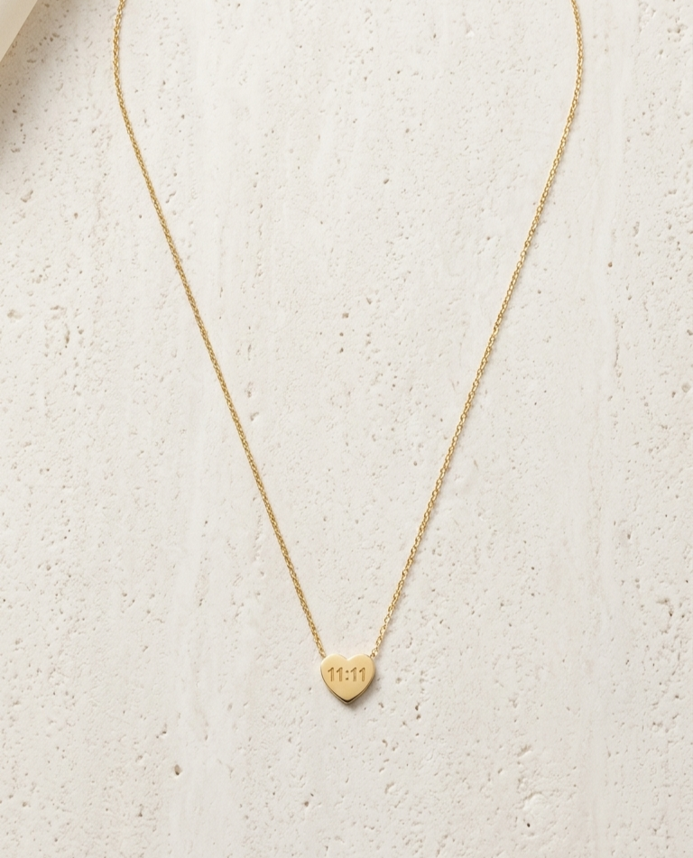 dainty gold-tone necklace featuring a small heart-shaped pendant engraved with the numbers "11:11," resting on a shimmering textured fabric.