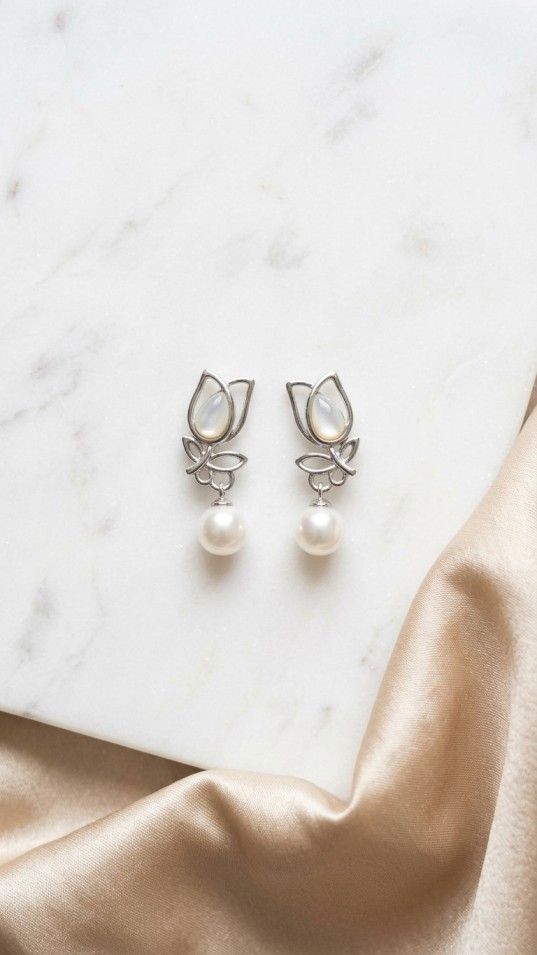 A pair of silver earrings shaped like stylized tulips with a luminous white inlay and a dangling round white pearl, resting on a white marble surface next to draped champagne silk.