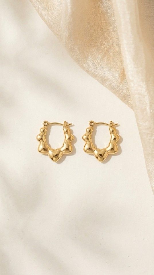 pair of small gold-tone huggie hoop earrings with a scalloped heart design, displayed on a shimmering white fabric background.