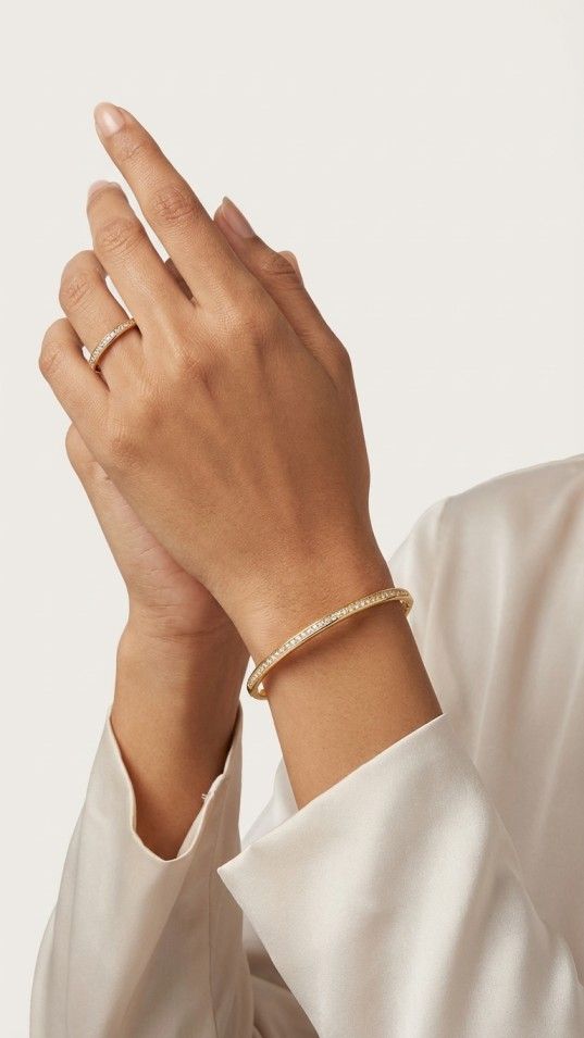 A minimalist gold-tone jewelry set featuring a thin circular bangle and a matching ring, both with a single row of small white crystals, placed on a silver-grey fabric.