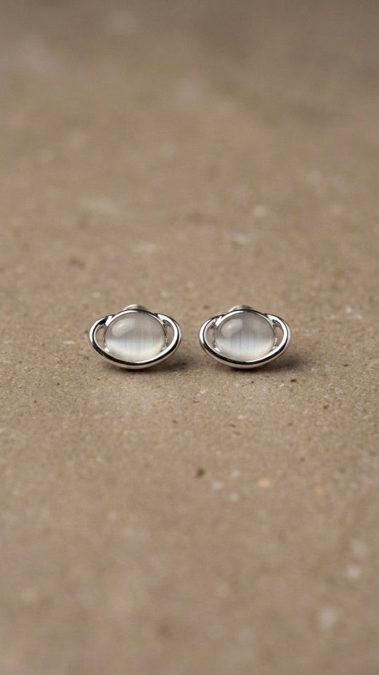 A pair of silver stud earrings shaped like the planet Saturn, featuring a luminous white cabochon stone in the center, resting on a textured beige surface.