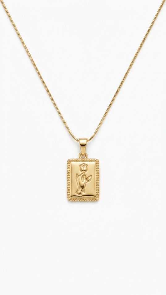 gold-tone necklace featuring a rectangular pendant with an embossed rose design and beaded border on a thin snake chain, resting on a shimmering white fabric background.