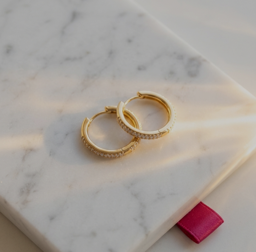 A pair of small gold-tone huggie hoop earrings adorned with a row of shimmering clear stones, resting on a white marble surface under soft sunlight.