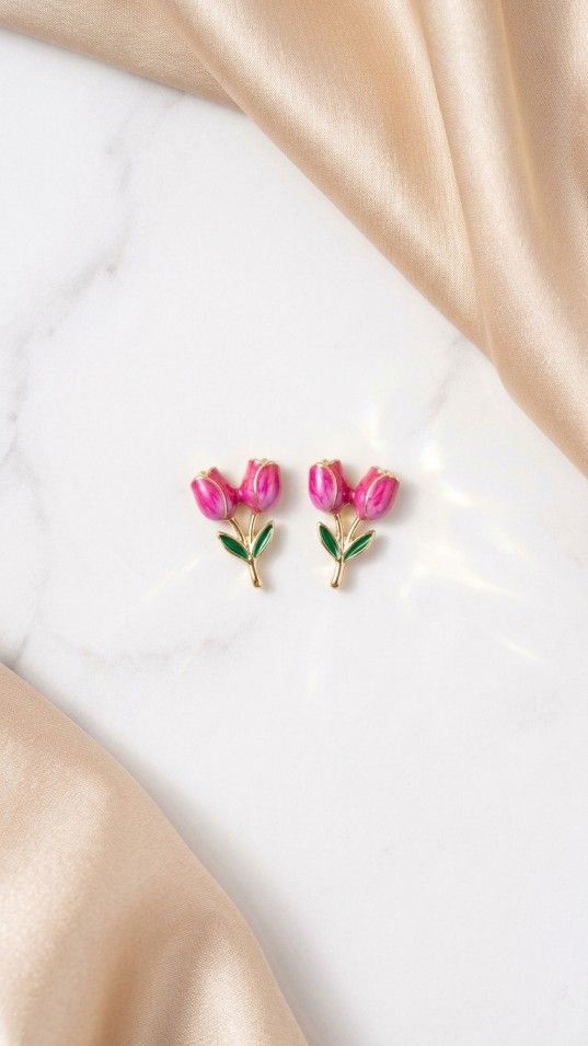 A pair of gold-toned stud earrings featuring two pink enamel tulip flower buds with green leaves, displayed on a white marble background with champagne silk fabric.