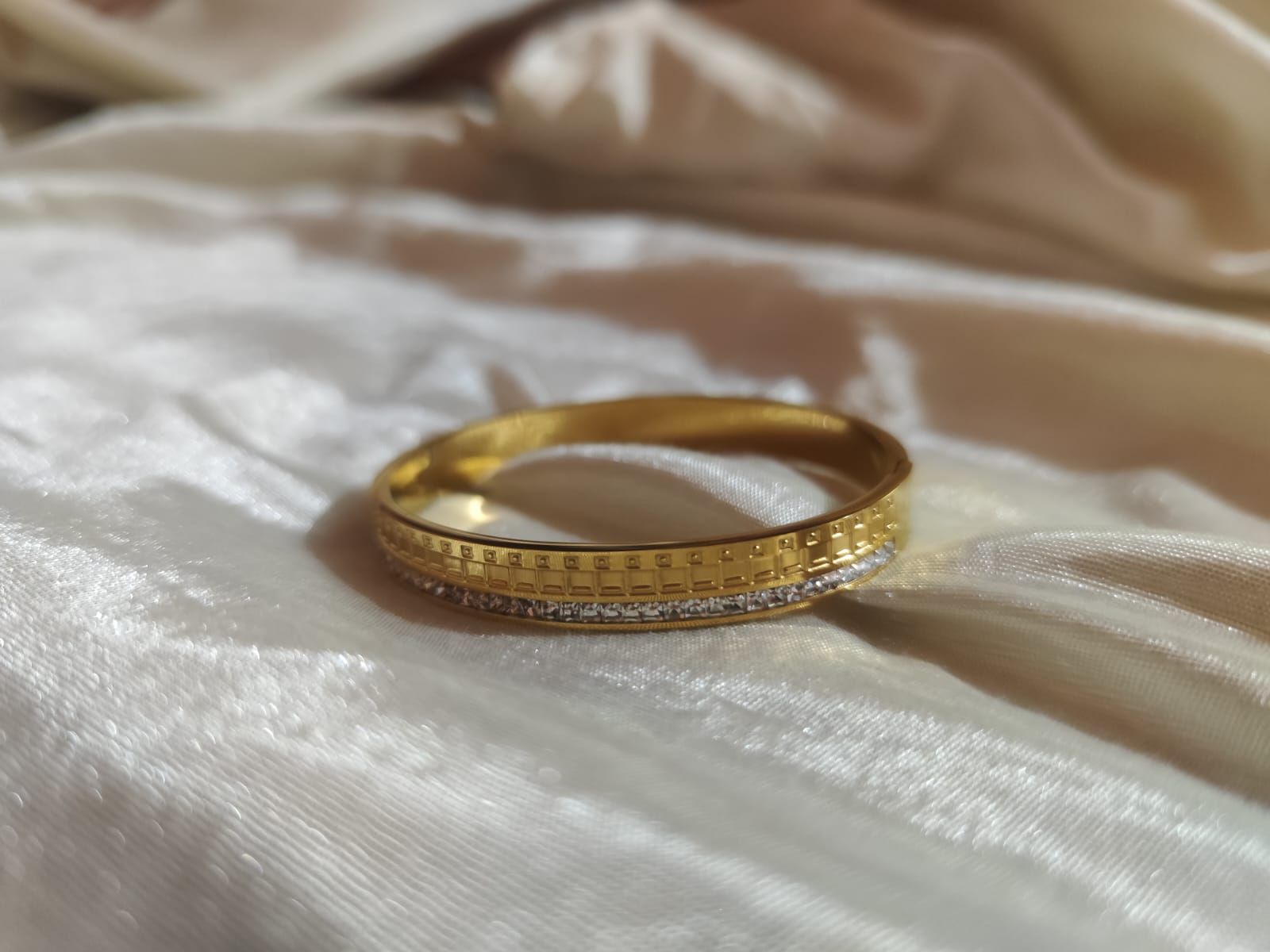 gold-tone bangle featuring a top half with an embossed square grid pattern and a bottom half lined with small white crystals, set against a white silk background.